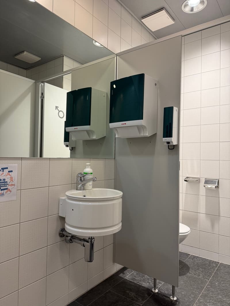 A white tiled bathroom with a sink, mirror, and toilet. Dispensers for hand soap and paper towels are mounted on the wall.