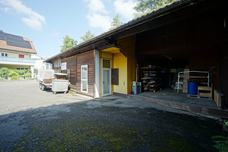 An old wooden house with a garage, yellow wall, white door, solar panels, barrels, shelves, trolley, and truck with container outside