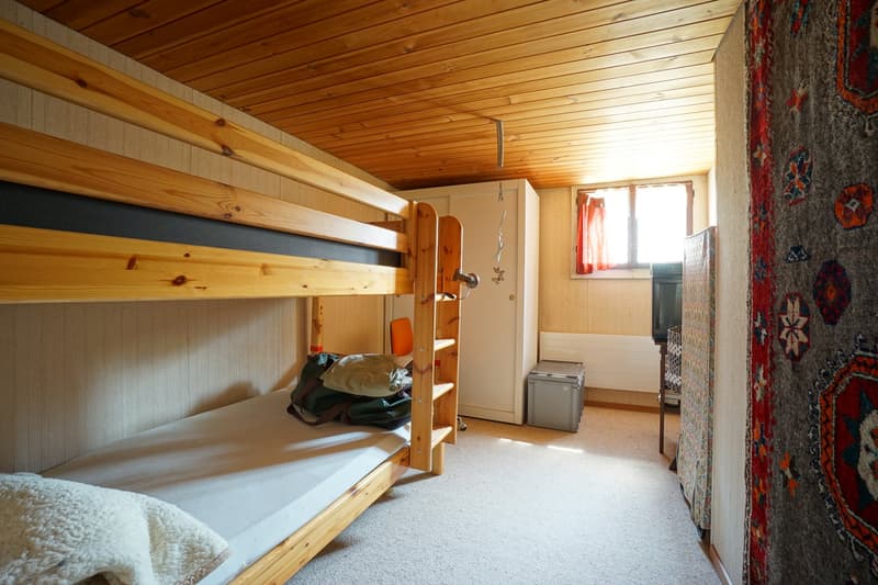 a room with two wooden bunk beds, white linen sheets, bags and pillows, wooden shelves, white walls, wooden ceiling, wardrobe, storage box, radiator and window with red curtains