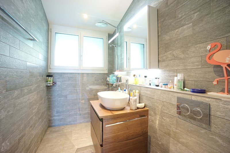 modern bathroom with gray tiles, white sink, wooden cabinet, shower area, mirror, and various toiletries