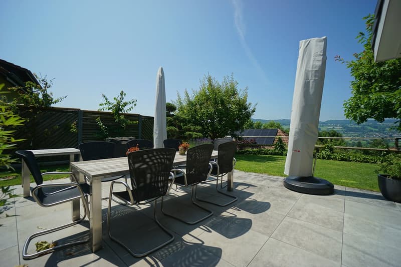 Outdoor dining table with chairs, covered by umbrellas, tiled patio, greenery, solar panels in the distance