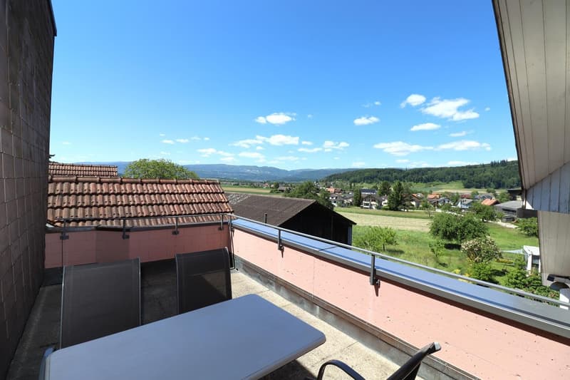 The image shows a balcony or terrace with a scenic view of the surrounding countryside. The balcony has a tiled roof and a glass railing, and there is a table and chairs on the balcony. The view beyond the balcony includes rolling hills, forests, and a vi