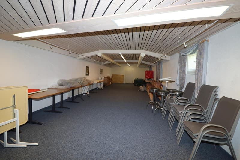 The image shows a large open space with a high ceiling and wooden paneled walls. The room is equipped with several long tables and stacks of chairs, suggesting it is used for meetings, events, or classes. The space appears to be well-lit with track lighti