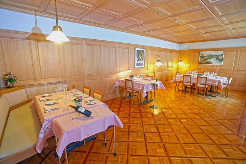 Spacious dining room with wooden paneled walls and ceiling, parquet flooring, multiple dining tables set with tablecloths and place settings, hanging light fixtures, and artwork on the walls.
