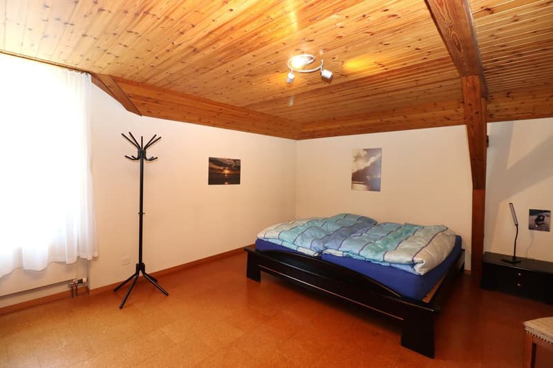 The image shows a bedroom with a wooden ceiling, a bed with a blue and green comforter, a coat rack, and two framed wall art pieces.