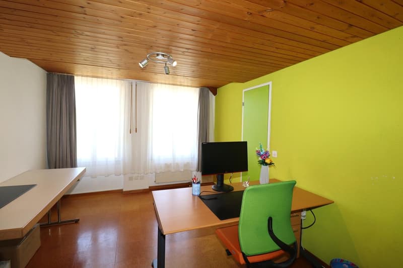 Spacious living room with wooden ceiling, large windows, desk with computer, green chair, and a fireplace
