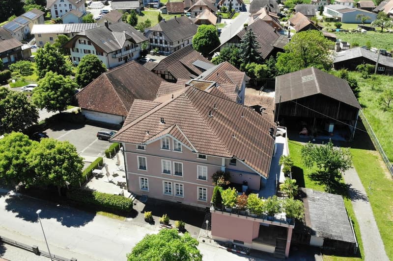 This image shows a multi-story building with a red tile roof surrounded by other buildings and trees in a small town or village setting. The building appears to have a parking area in front of it.