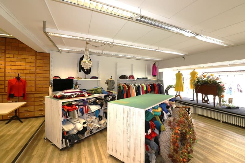 Large open retail space with clothing racks, shelves, and display items. Wooden floors, white walls, and overhead lighting. A central table with various items and a chandelier hanging from the ceiling. Colorful clothing and accessories are displayed throu