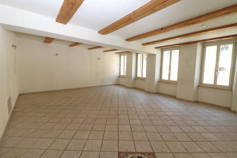 Large open space with exposed wooden beams on the ceiling, tiled flooring, and multiple windows providing natural light.