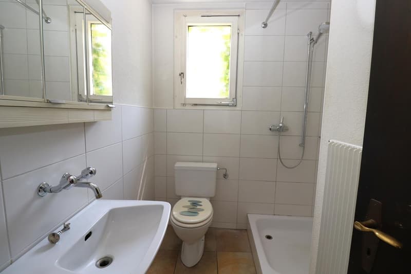 The image shows a bathroom with white tiled walls, a toilet, a sink, and a shower area with a window. The bathroom appears to be clean and well-maintained.