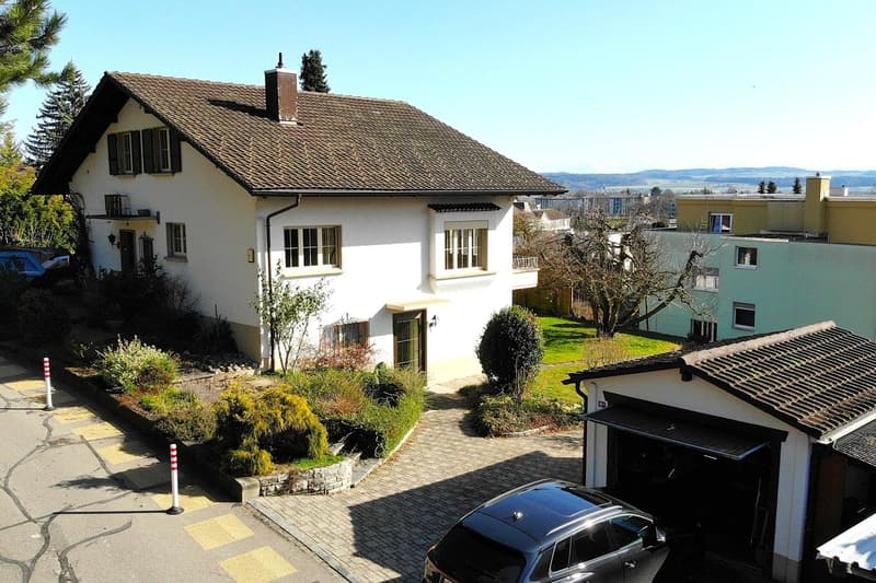 white house with brown roof, two-story, black shutters, brick driveway, car parked in front, surrounded by plants, trees and grass, view of mountain in distance