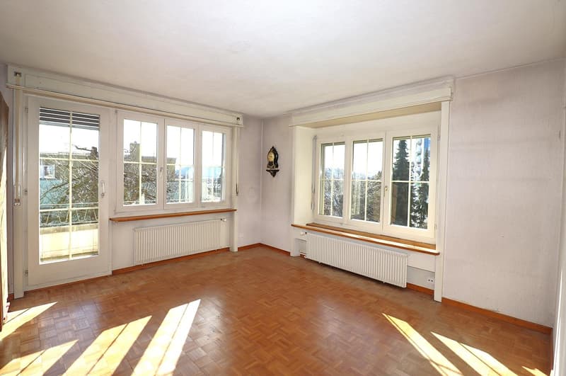 Empty room with wooden floors, two sets of glass sliding doors leading to a balcony, white walls, two large windows, white radiators, wooden window sills, view of the outside with trees