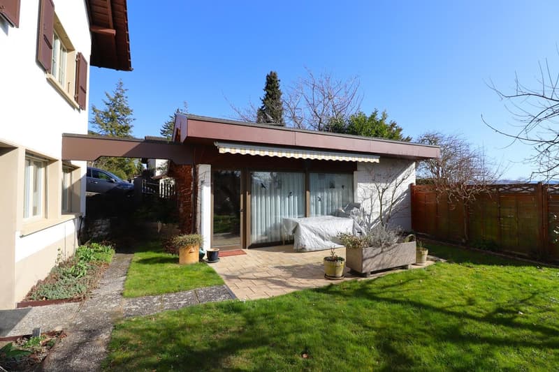 Detached house with awning, terrace, and covered parking area. Garden with brick pathways, green grass, and potted plants.