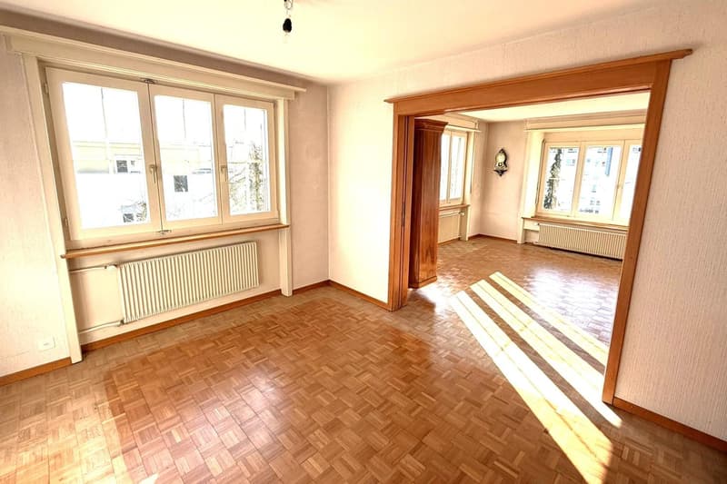 large room, wooden floors, white walls, three windows with white frames, double doors to another room, radiators below the windows
