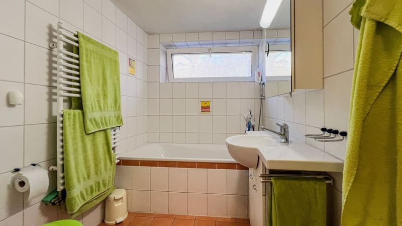 Bathroom features a white bathtub, white sink, and tile walls. There is a towel rack with green towels. There is a white toilet and a toilet paper holder.