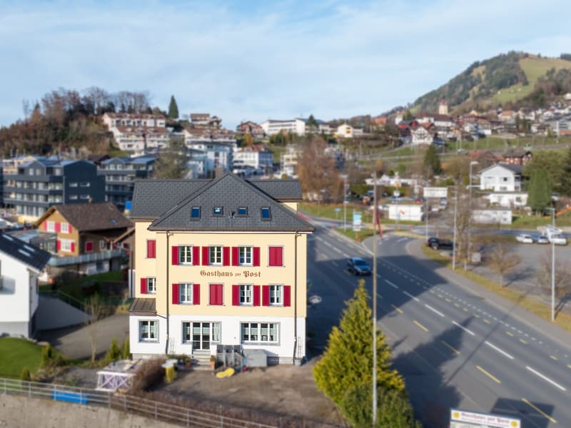 Aerial view of Gasthaus zur Post, a building with a slate roof and red shutters, situated in an urban setting with surrounding houses and trees.