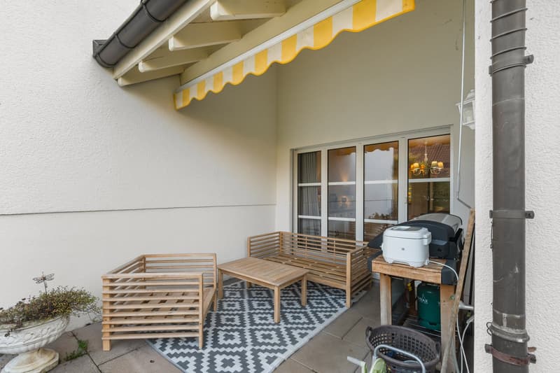 A covered balcony area with wooden furniture, a rug, and a barbecue.