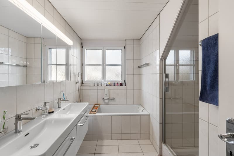Modern white tiled bathroom with bathtub, glass door shower, double sink, mirror and two windows