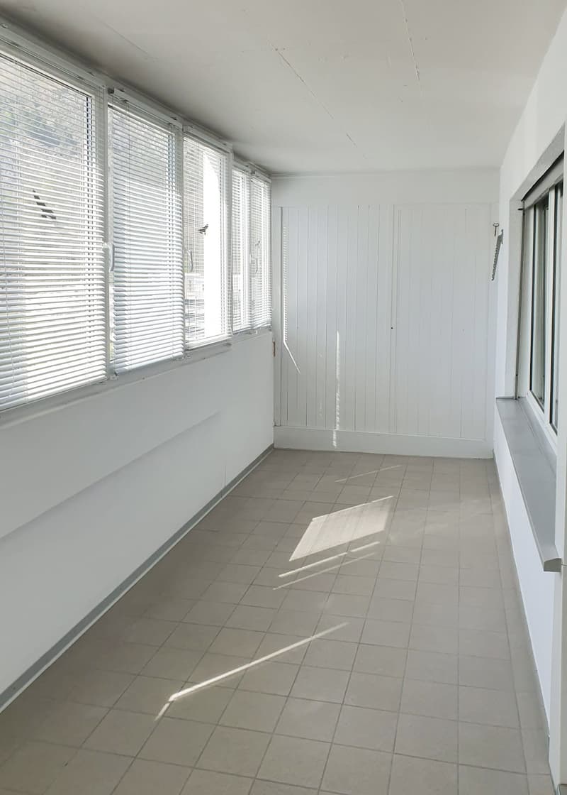 Empty room with white walls, tiled floor, large windows with blinds, and a door.