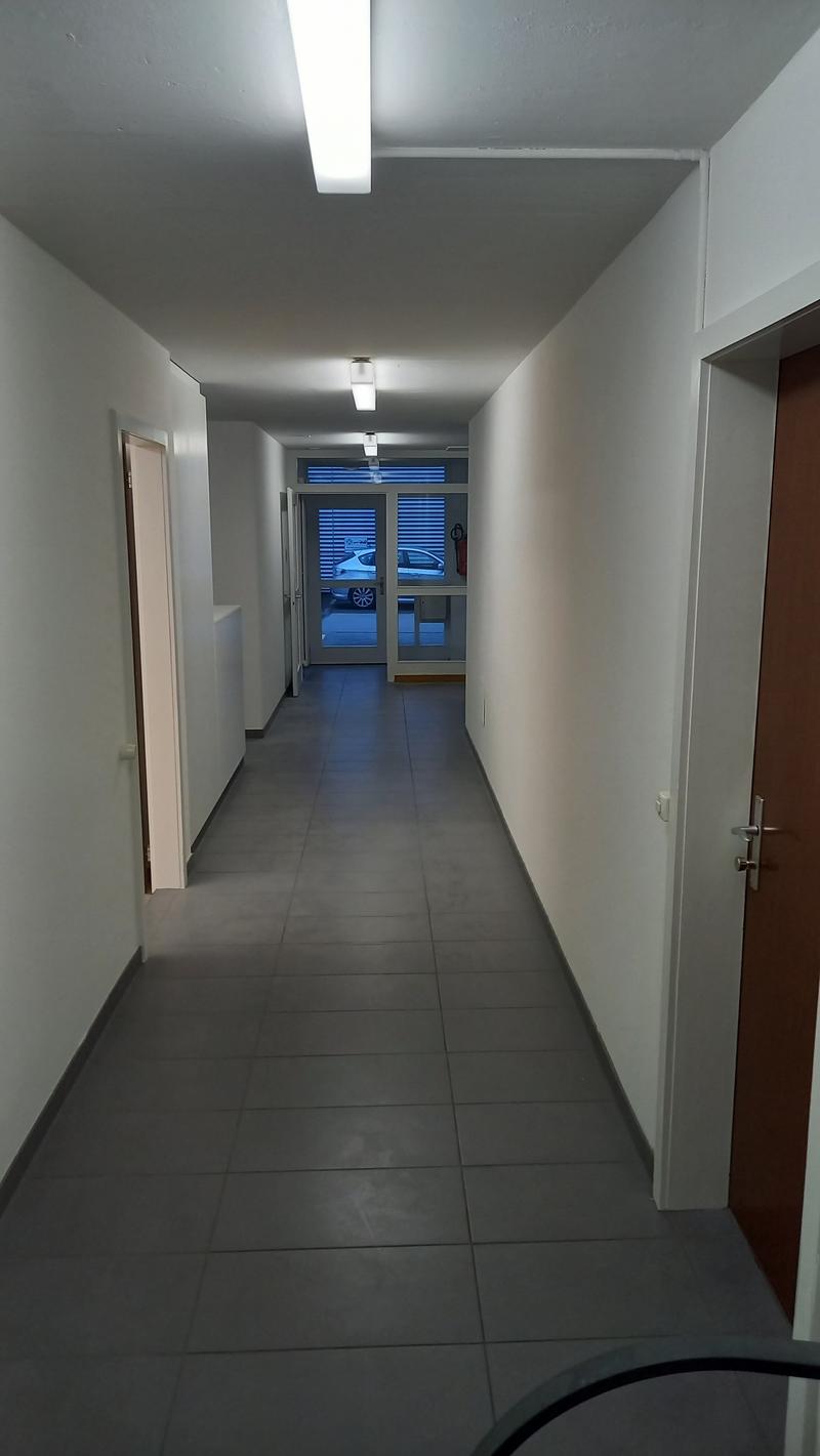 The image shows a long, narrow hallway with gray tile flooring and white walls. The hallway has recessed lighting fixtures in the ceiling and several doors leading to other rooms. Through the glass door at the end of the hallway, a car can be seen parked