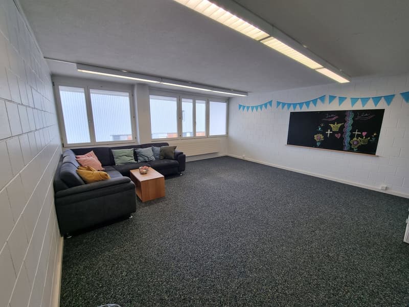 Large room with white walls, gray carpeted floor, and several windows. There is a black chalkboard on the wall and colorful bunting decorations. The room contains a large black sofa and a wooden coffee table.