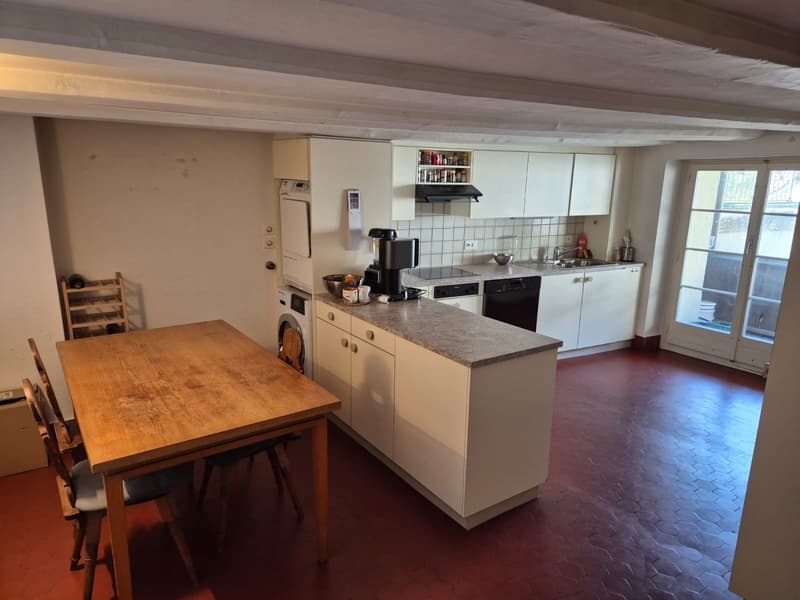 Spacious kitchen with white cabinets, granite countertops, and appliances including a dishwasher. There is a wooden dining table with chairs. The kitchen opens up to a living area with tile flooring.