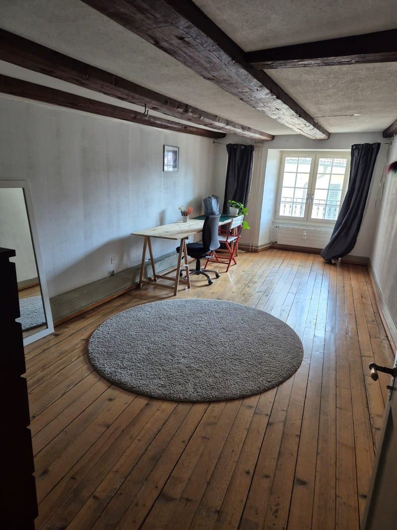 Spacious room with wooden beams on the ceiling, hardwood floors, a large round rug, a desk with a chair, and a window providing natural light.