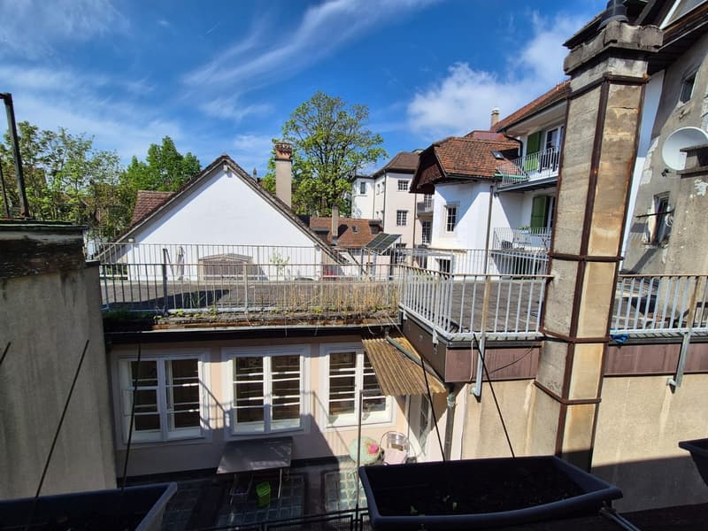 Multi-story building with white walls, red tile roofs, and balconies. Trees and greenery visible in the background. The building appears to be in a residential or historic area.