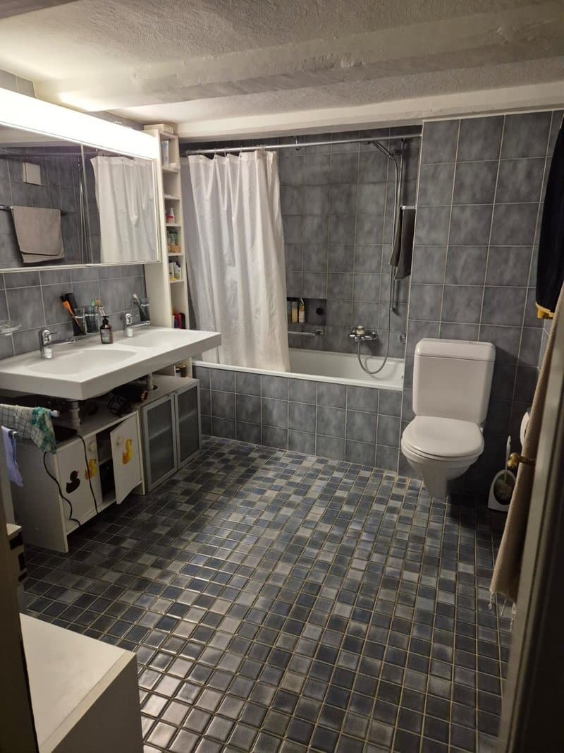 Spacious bathroom with gray tile flooring, white vanity with sink, toilet, and a bathtub with a shower curtain