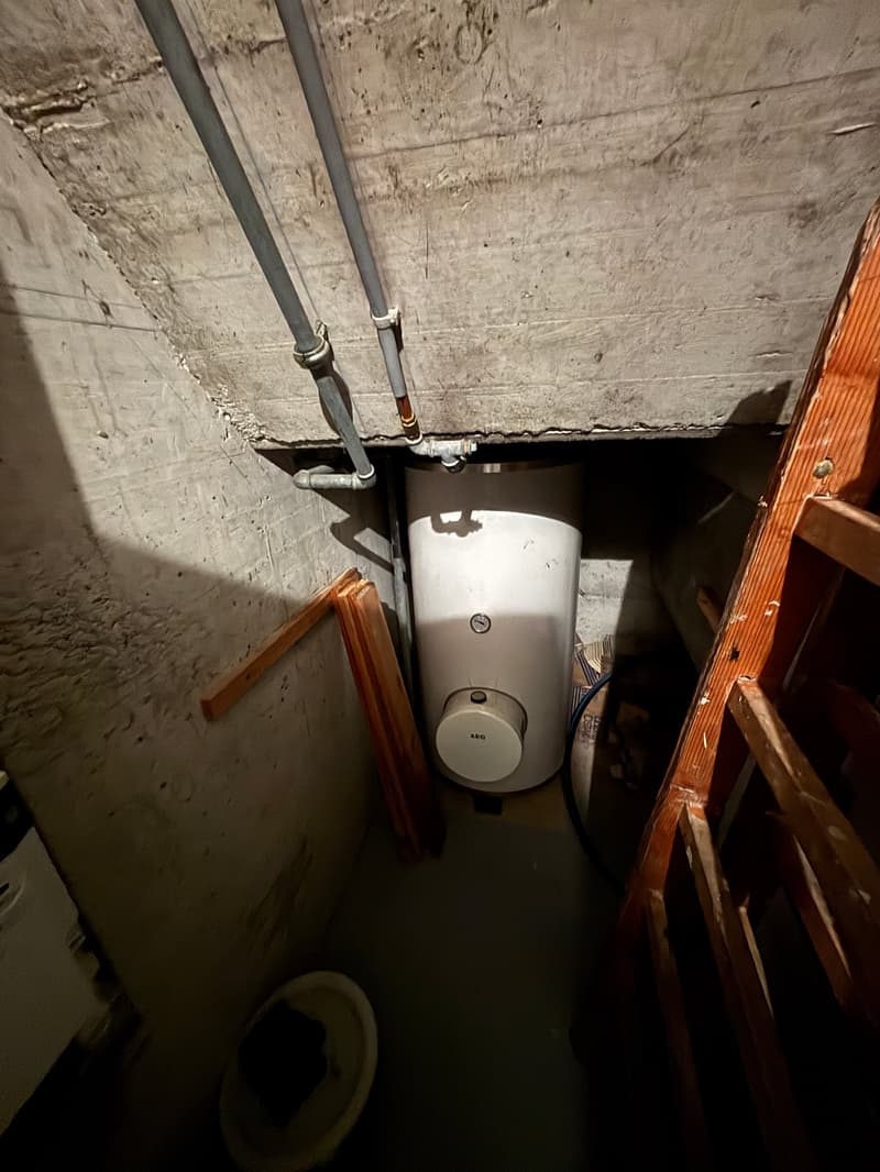 A small room with a white water heater, wooden ladder, and pipes attached to the ceiling.
