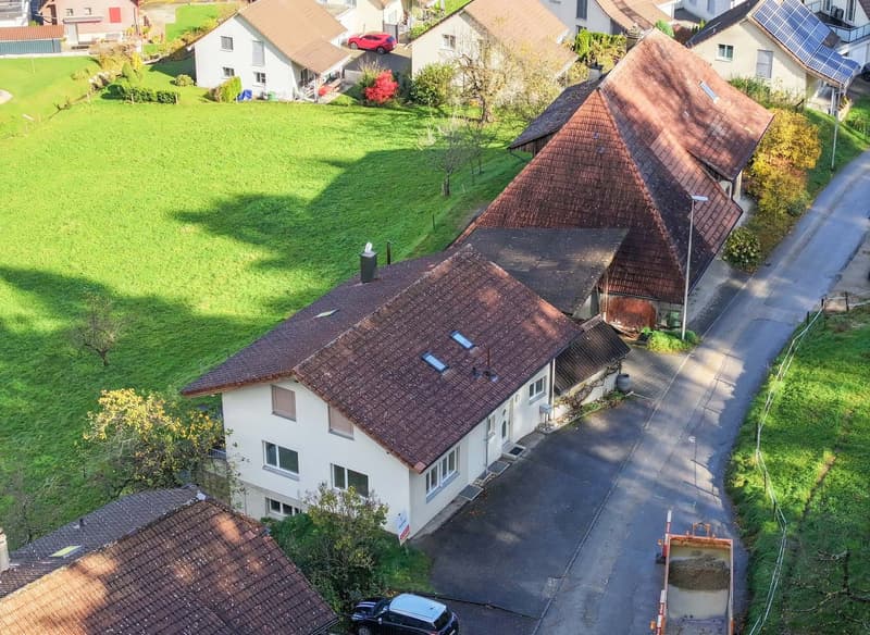 Aerial view of a row of houses with brown roofs and white walls, surrounded by green grass and trees, with a driveway and a parked car in front.