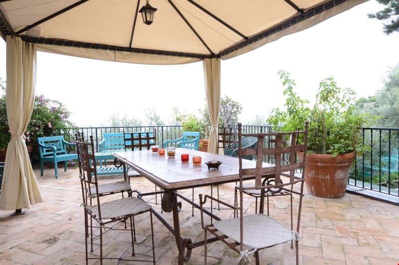 Covered outdoor dining area with a large wooden table and wrought iron chairs, surrounded by potted plants and overlooking a scenic garden view. The space is shaded by a beige canopy and features a fireplace for ambiance.