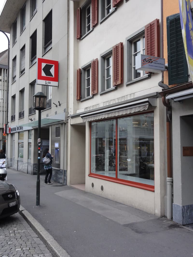 A multi-story building with white walls, red shutters, and large windows. There is a red directional sign on the building and a storefront with large display windows.