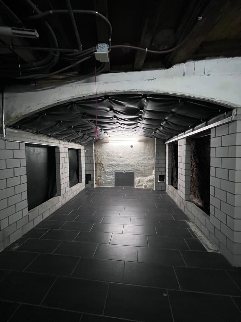 Empty room with black tiled floor, white brick walls, black curtains, and exposed ceiling with pipes and lights