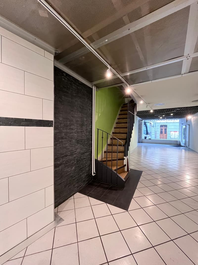 Empty hallway, white tiles, black mat, black and green wall, wooden stairs, metal railing, lights on ceiling