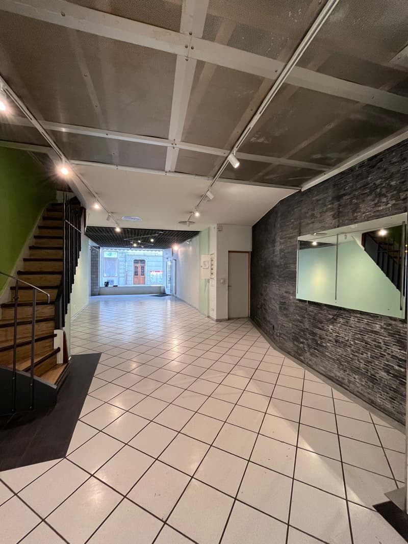 Empty room with white tiles, wooden staircase, brick wall, mirror, and lights on the ceiling.