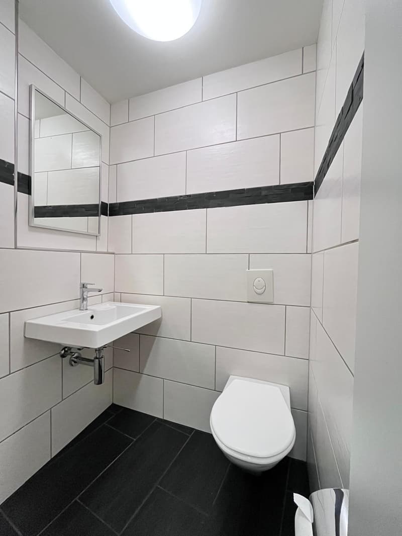 small bathroom with white tiles, black tiles on the floor, white sink, silver faucet, white toilet, mirror