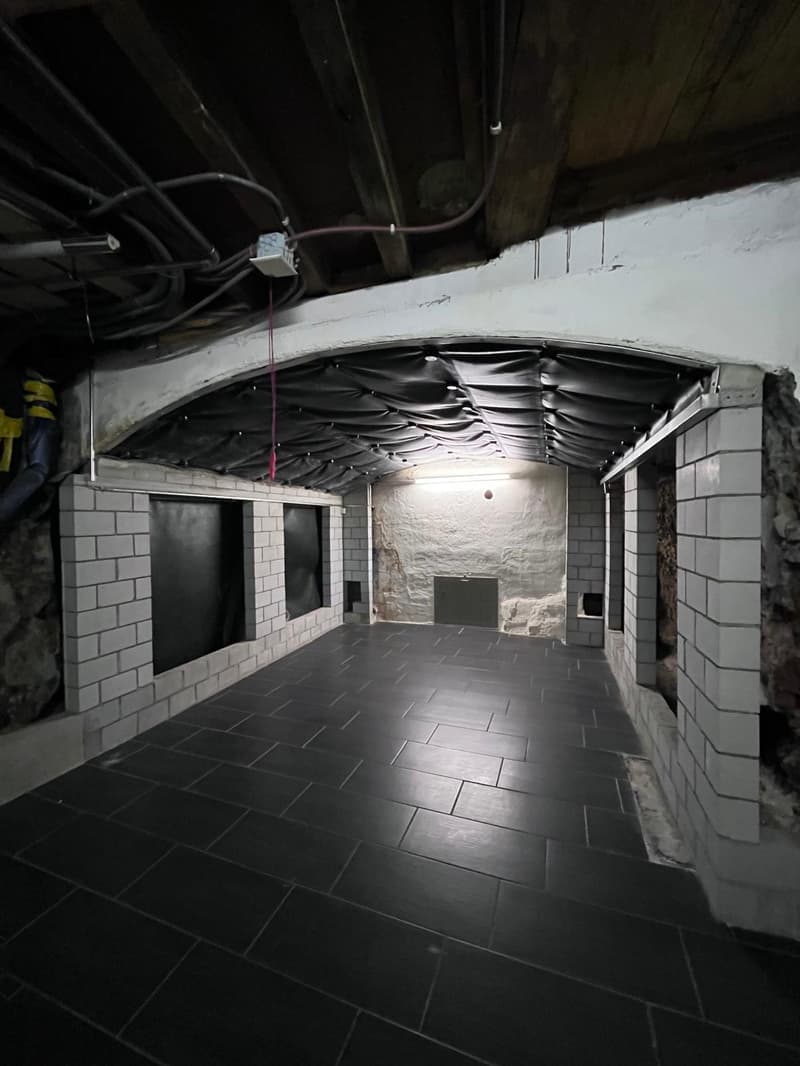 Empty basement with black tiled floor, white brick walls, black doors, and exposed ceiling with pipes and wires.