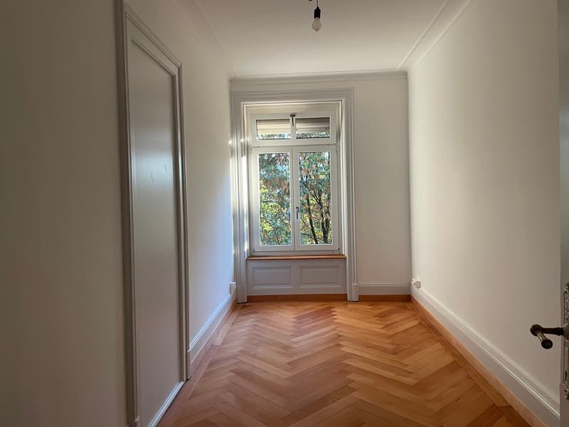 Empty room with parquet floor, white walls, central light, and large window with view of outside