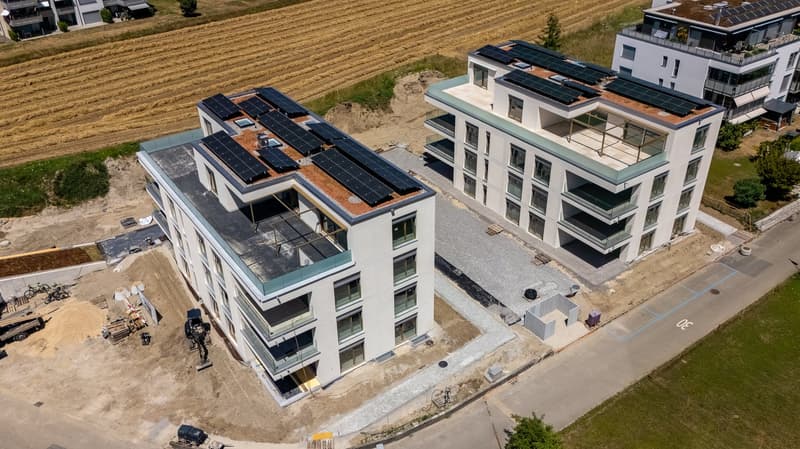 Multi-story apartment building with solar panels installed on the roof, surrounded by a grassy field and construction site
