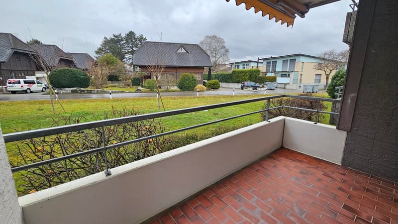Balcony with metal railing, red brick floor, view of a grassy field and houses