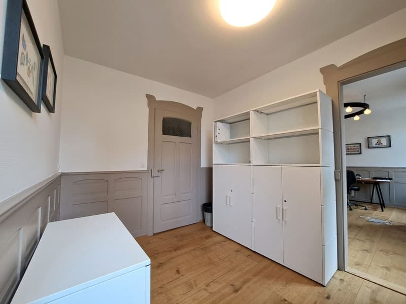 Empty room with wooden floor, white walls, white cabinets, door, trash bin, framed pictures on the wall, ceiling light, and a mirror.