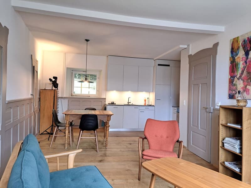 Open-plan living and dining area with wooden floor, white kitchen cabinets, dining table, chairs, wooden coffee table, armchair, sofa, wooden bookshelf, pendant light, and a window.