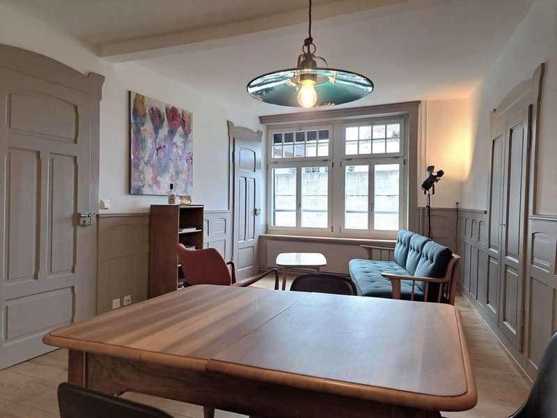 A room with a wooden table, a couch, a chair, a shelf, a lamp, a painting, and a window.