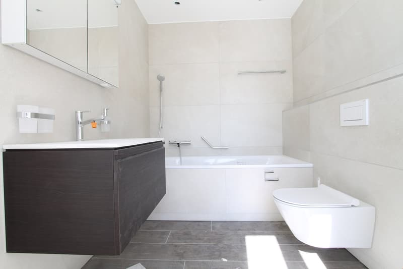 Large bathroom with white walls, gray tile flooring, a freestanding bathtub, a vanity with a dark wood cabinet, and a wall-mounted toilet.
