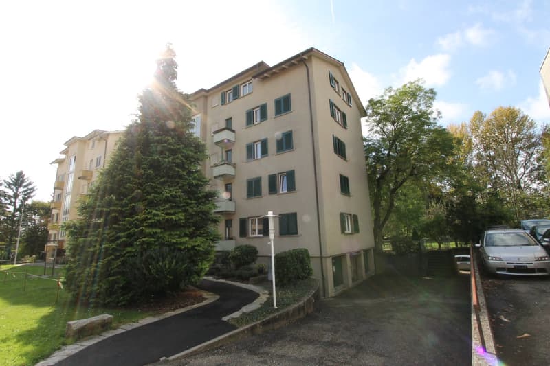 4 story building with beige color, multiple balconies, green trees, paved walkway, cars parked, pathway.