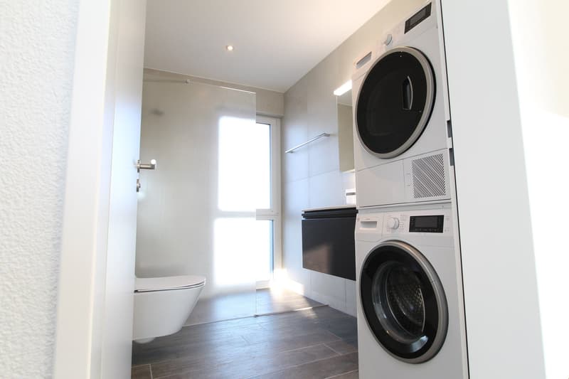 modern bathroom with white tiles, white toilet, glass shower, washing machine, and dishwasher