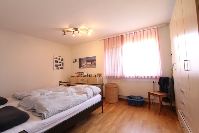 Bedroom with bed, wooden floor, wardrobe, window with curtains, small table, basket, and a radiator.