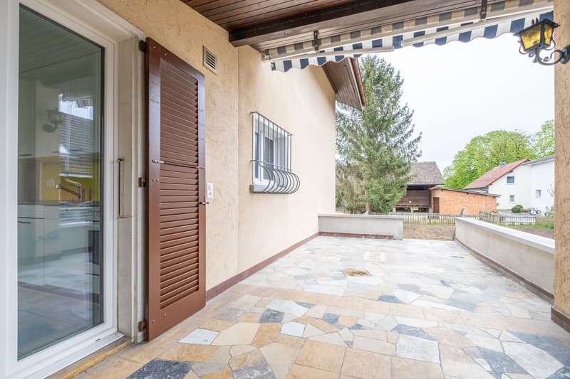 patio with stone floor, brown wooden door, glass door, awning, wall with window, outdoor light
