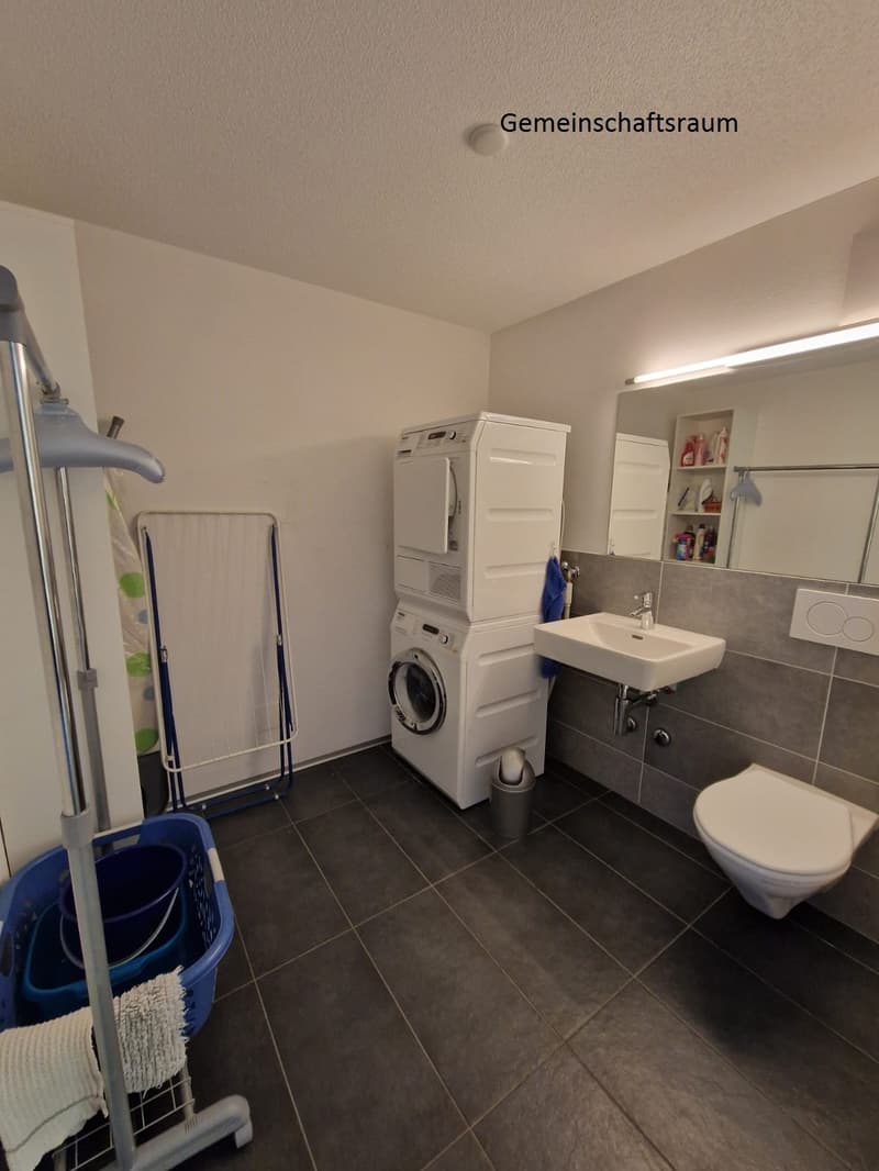 White walls, grey tiles on the floor, stackable washer and dryer, sink, mirror, toilet, washing machine basket, clothes drying rack